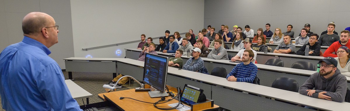 Banner image: shows professor standing at front of class, facing students sitting in rows