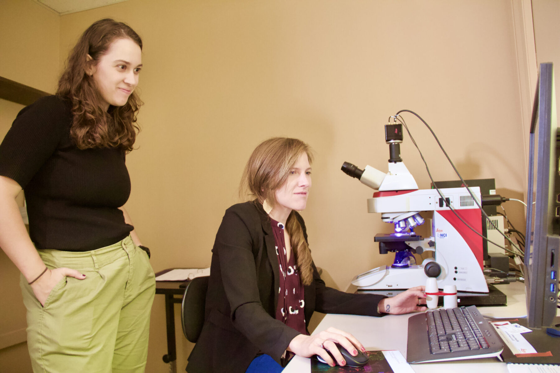 Professor Sydney Trask sits at her digital microscope while graduate student Erisa Met Hoxha stands and observes.