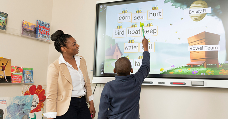 A young boy points out words in a reading game on a large screen, with a clinician standing next to him.