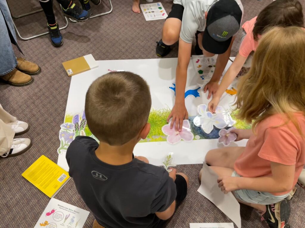 Children placing paper cutouts of flowers & plants on a large piece of illustrated paper on the floor.