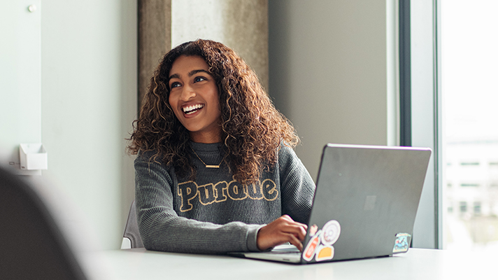A female college student wearing a Purdue shirt, sitting in front of a laptop and smiling.