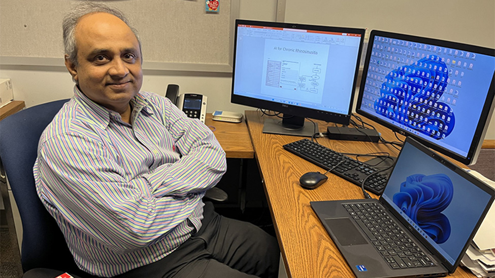 College professor sitting in front of a laptop and two computer screens smiling at camera.