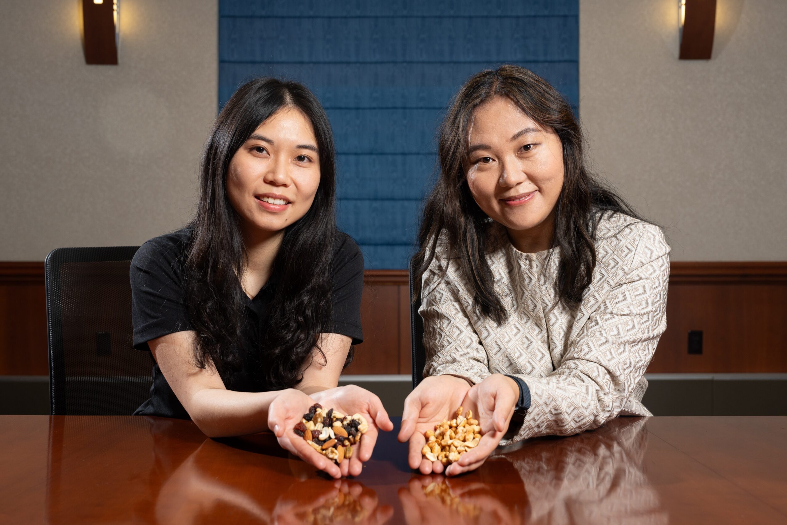 Two researchers seated at a conference table smile at the camera while holding nuts and trail mix.