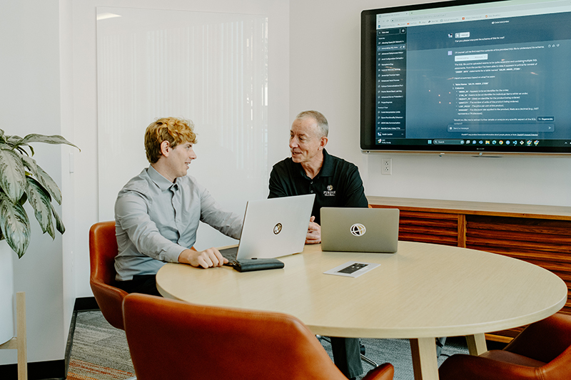 Austin Lawton and Jon Harbor chat at a table with two laptops open and a large screen on the wall behind them.