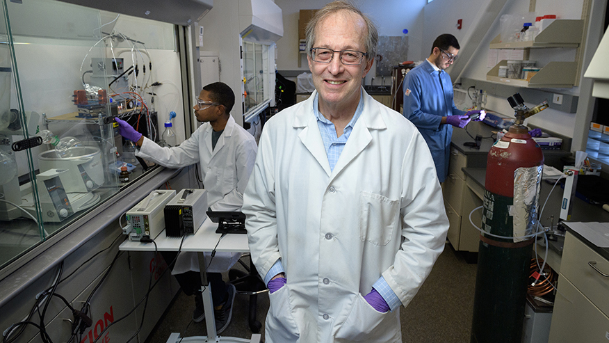 A Purdue researcher in a white lab coat stands in his lab as two other scientists conduct research behind him.