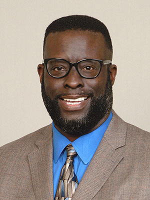 Man wearing glasses, smiling in a tan paid suit with a blue shirt.