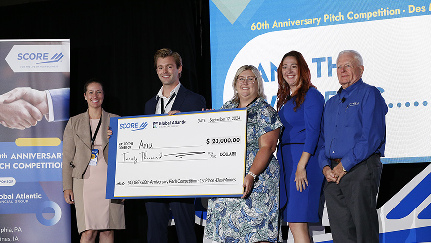 Three women and two men in business attire stand together. One man holds a $20,000 check written to Anu.