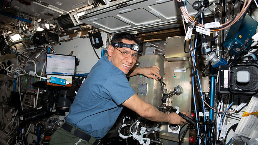 A man wearing a headlamp floats while adjusting gauges on equipment encased in shiny, steel-looking rectangular compartments