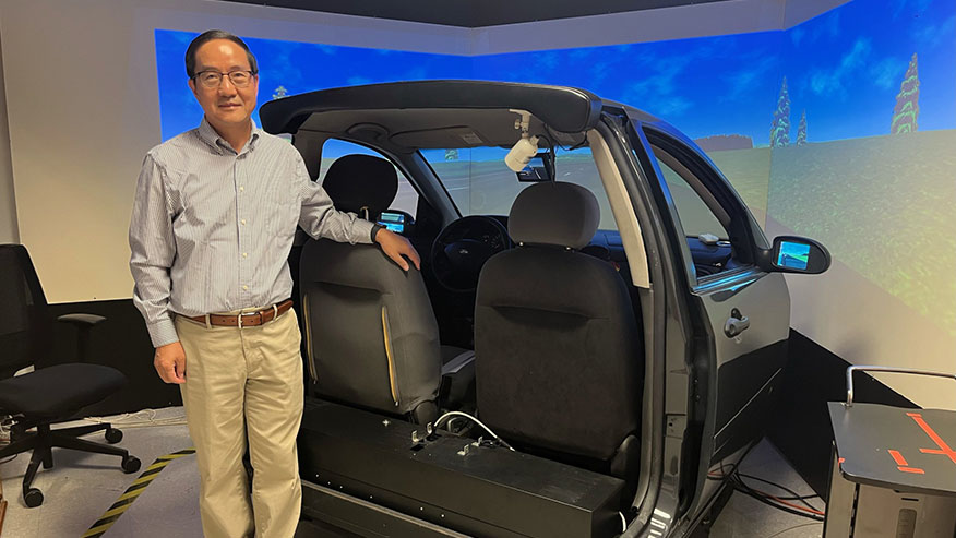 A Purdue professor stands behind the seats of a driving simulator.