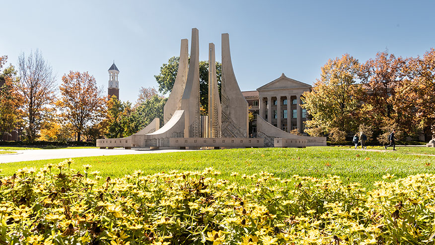 Wide shot of the Class of 1939 Water Sculpture, Hovde Hall and the Bell Tower with yellow flowers in the foreground.