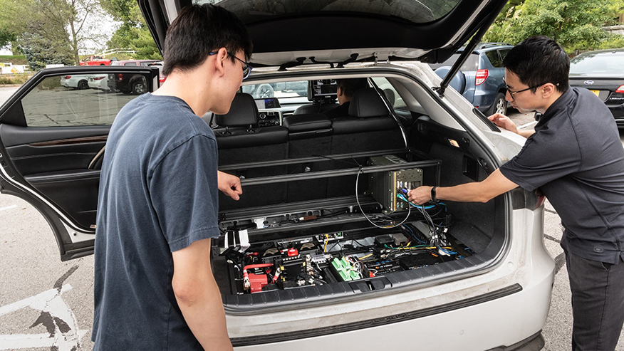 A student and professor on either side of a vehicle’s open trunk look at electronic devices and wiring installed inside