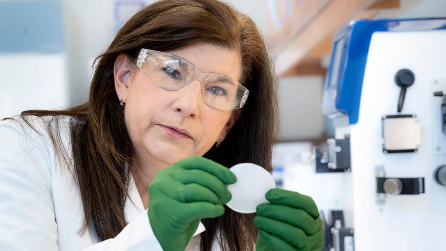 Researcher wearing goggles, white lab coat and green gloves closely looks at a white circle she holds in her hands.