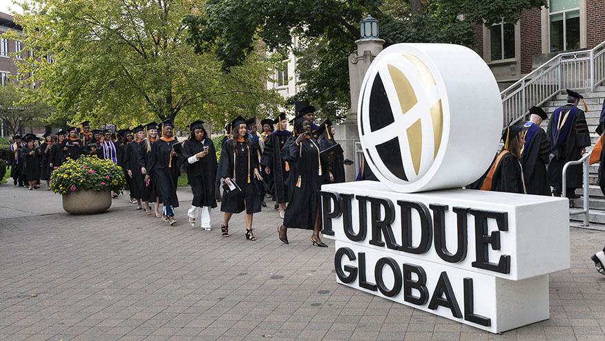 Purdue Global students in caps and gowns walk down a sidewalk past a large Purdue Global logo.