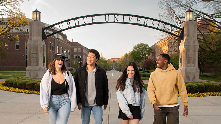 Four smiling college students walking on Purdue University's campus under the Gateway to the Future arch.