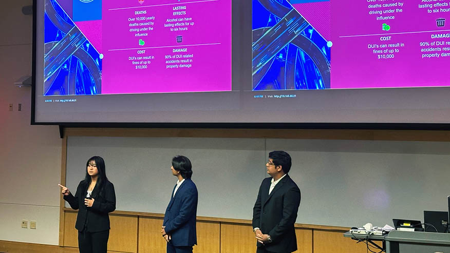 Three Purdue students make a business pitch to judges while standing under a video screen that shows purple slides.