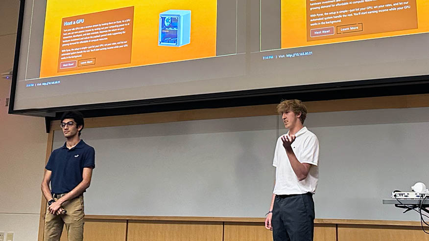 Two Purdue students make a business pitch to judges while standing under a video screen that shows yellow slides.