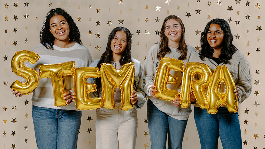 Four young women hold balloons that show the message STEM Era.