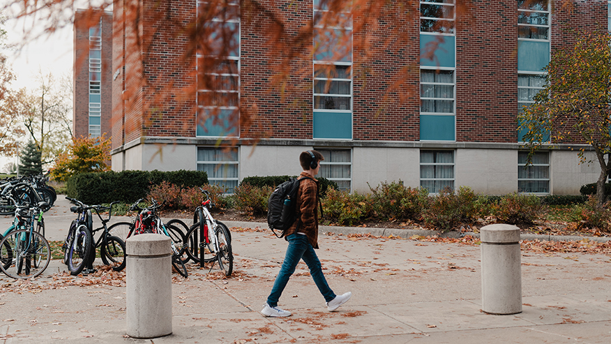 A student wearing headphones walks past one of Purdue University’s residence halls on a fall day.