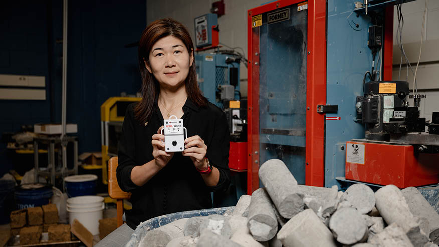 Wavelogix founder and Purdue researcher Luna Lu holds a REBEL sensor as she sits behind a container filled with concrete cylinders.