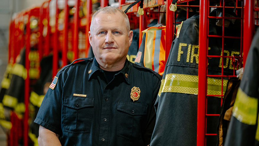 Purdue Global graduate and firefighter Michael Pruitt poses next to a row of turnout gear in a fire station.