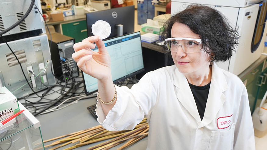 A Purdue researcher wears goggles and a white lab coat in a laboratory and holds a small, white circle to the camera.