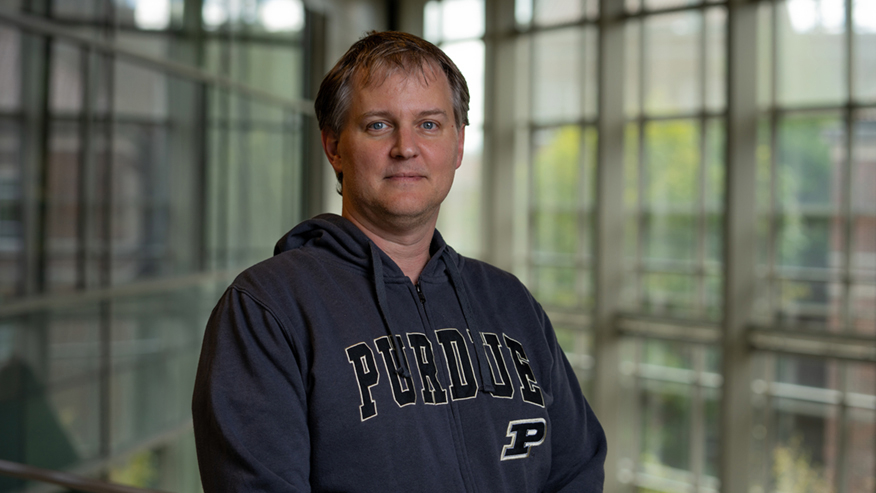 A Purdue University researcher wearing a black Purdue sweatshirt poses in an atrium.