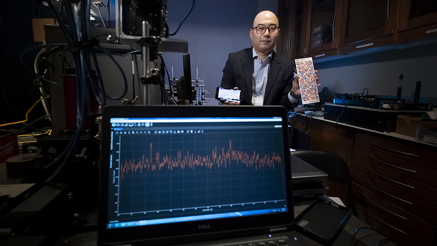 A Purdue researcher holds a smartphone and a color grid while he stands behind a computer screen that displays a line graph.