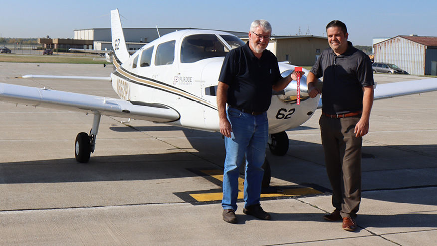 Two Purdue inventors stand in front of a Piper Archer aircraft at Purdue University Airport. One holds a red safety guard.