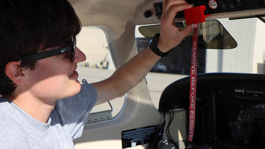 A Purdue student pilot wearing sunglasses adjusts a red safety guard clipped over a switch in an airplane’s flight deck.