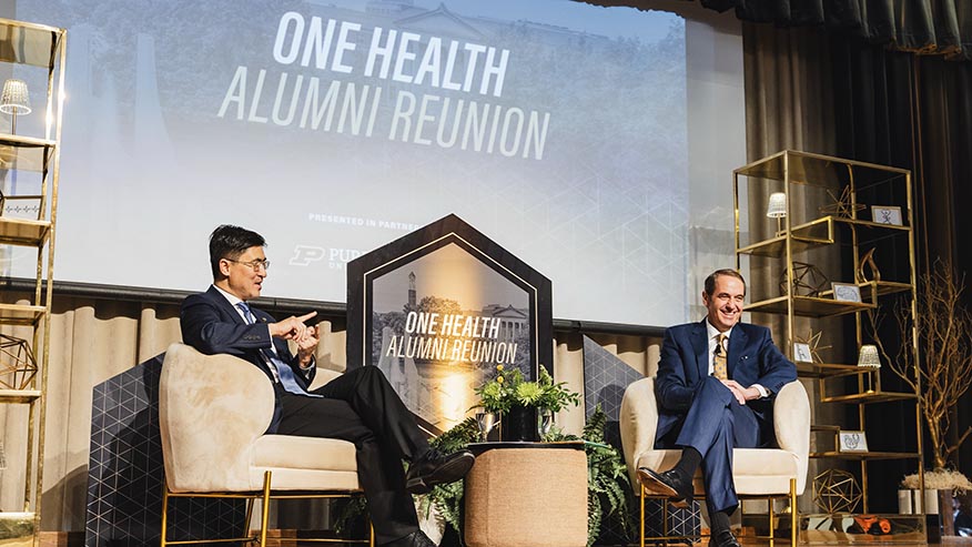 Two men sitting in chairs talk in front of a screen that reads, One Health Alumni Reunion.