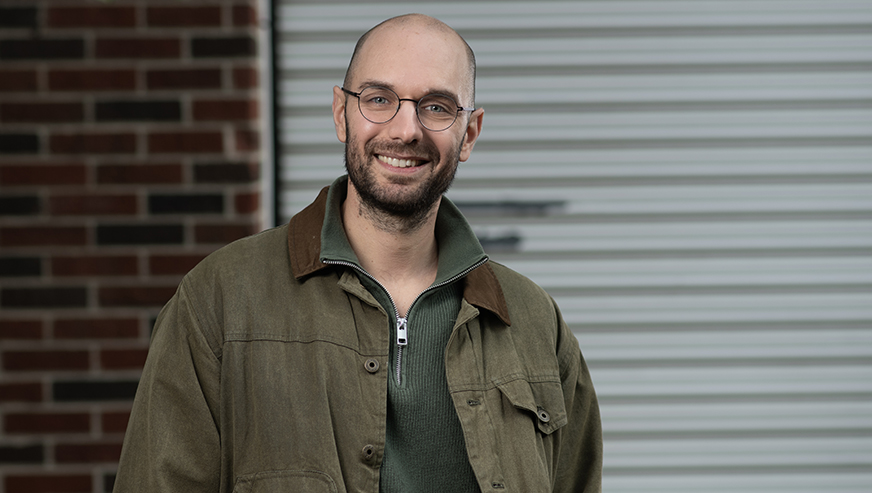 Alex Psomas smiles in front of a brick wall and metal garage door.