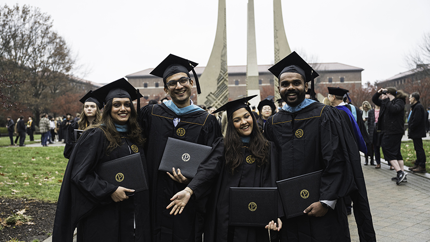 Four students graduating from Purdue University pose with their diplomas in front of the fountain on the Purdue Mall.