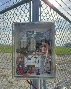 The SAFE-RWSL system mounted to a fence post; a box contains a camera lens and wires of different colors and thicknesses.