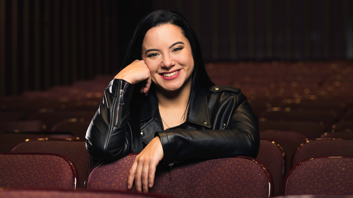 A woman sitting in a theater row rests both arms on the back of the seat in front of her.