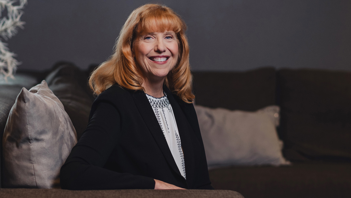 Jody DeKorte sits on a couch, half-turned toward the camera with one elbow on the armrest.