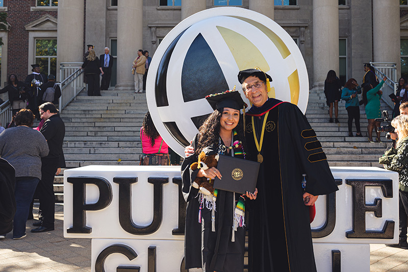 Purdue Global Chancellor Frank Dooley poses with a graduating student in front of a large Purdue Global logo.