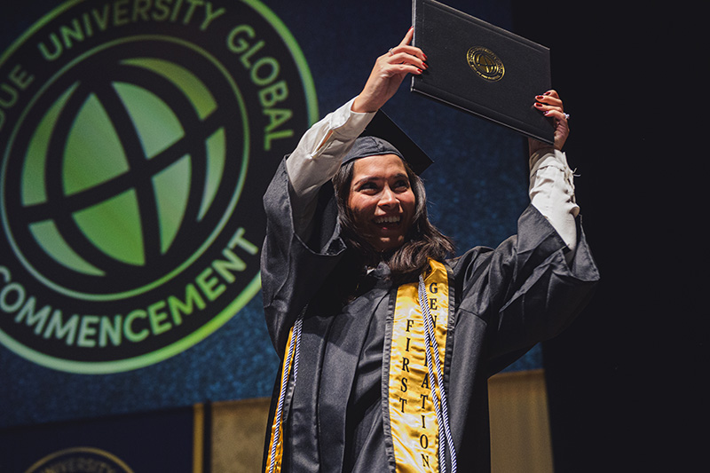 A Purdue Global graduate holds her diploma over her head in celebration as she walks across the stage.