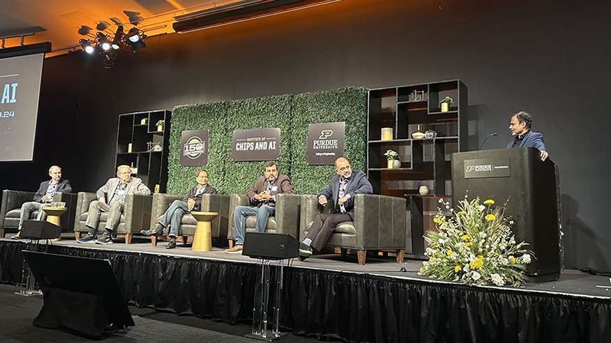 Kaushik Roy speaking from behind a lectern with a Purdue logo while four men in chairs on the left side of the stage turn toward him