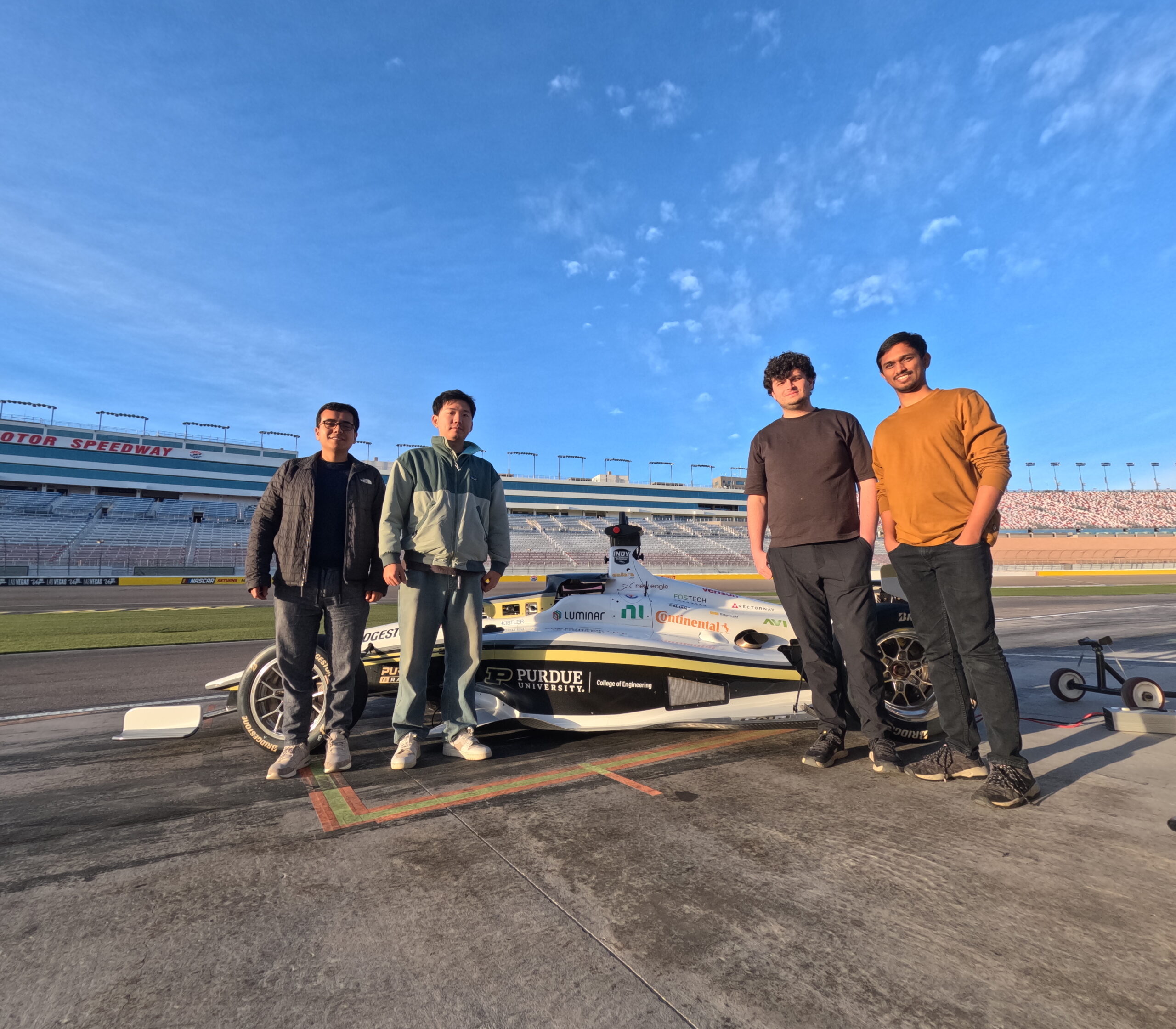 Four Purdue AI Racing team members stand in front of a race vehicle on a track within a speedway