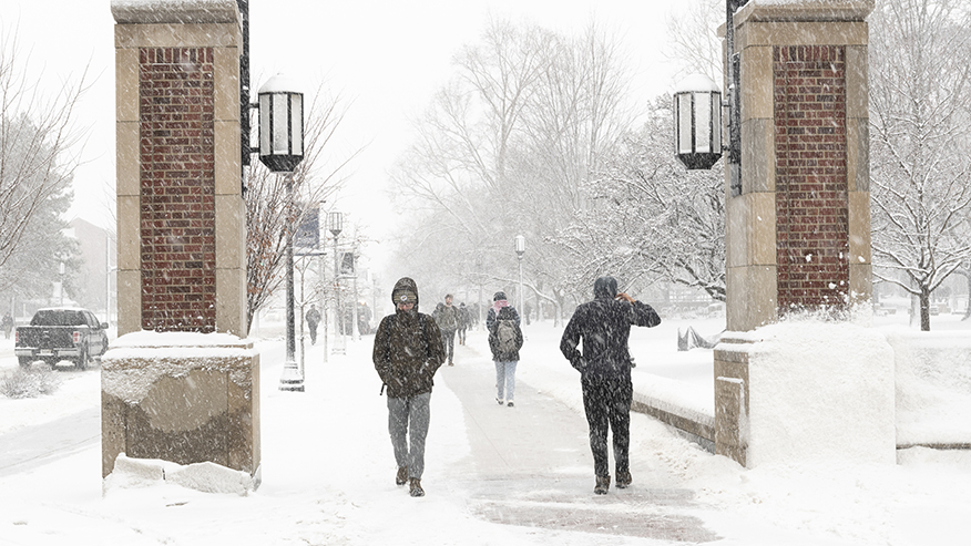 Students walk between brick pillars on a snowy day at Purdue University.