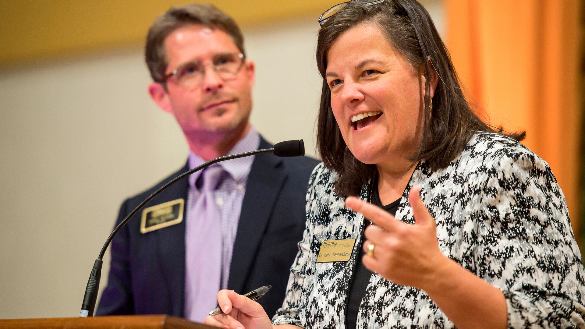 Dr. Katie giving a presentation with Todd Wetzel looking on in the background. 