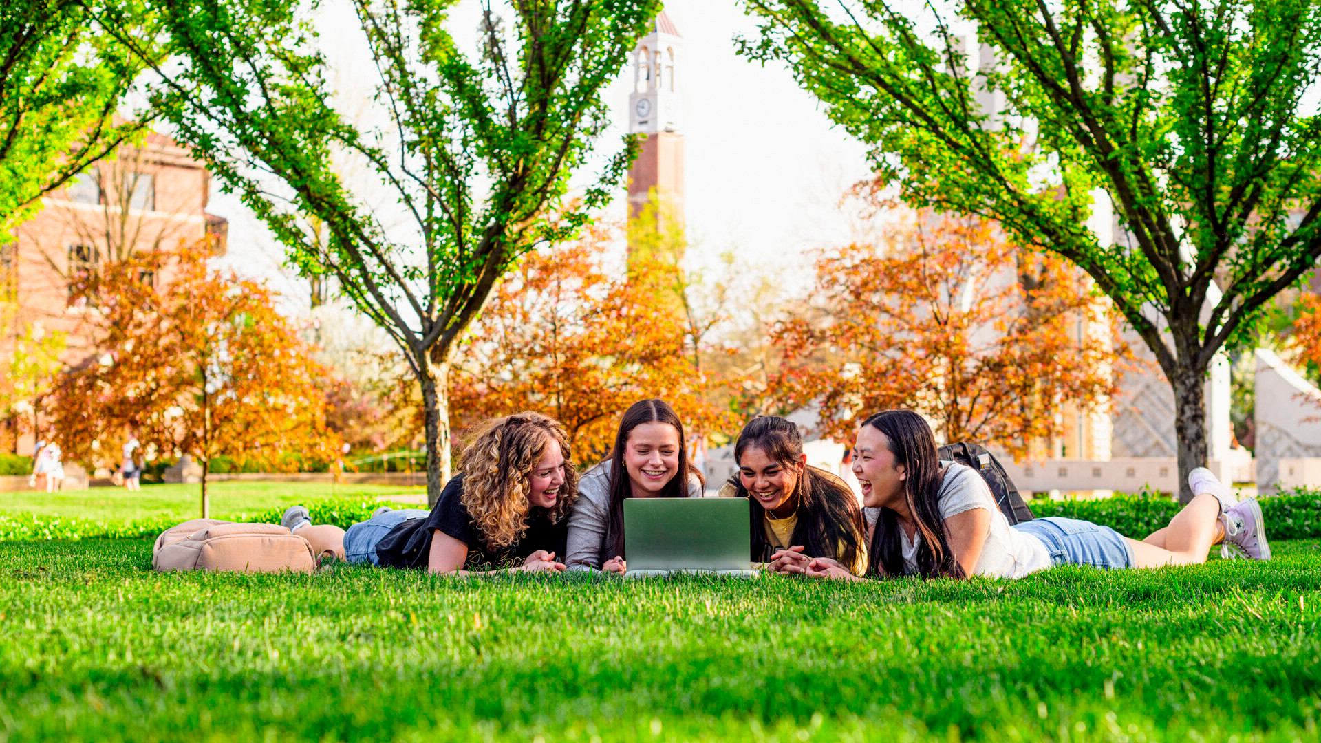 Four students laying in the grass in the green space in front of the Bell Tower and Engineering Fountain. 