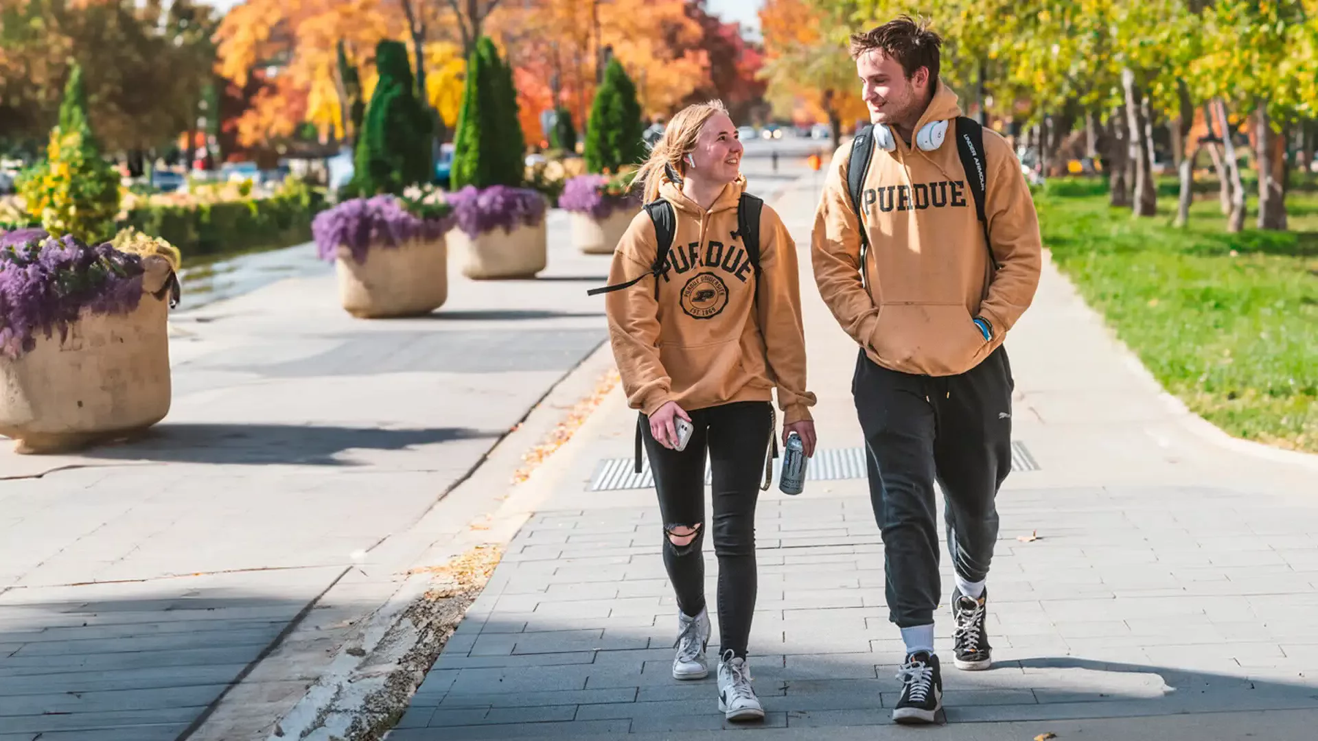 Two students walking and talking on campus in spring, wearing Purdue gear and smiling at each other.