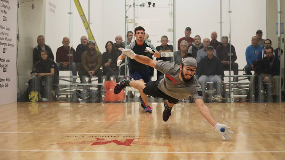 pictured: two men play four wall handball, one man is diving to hit the ball